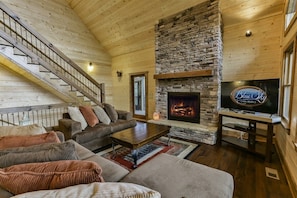 A cozy cabin interior with a stone fireplace, a television, and a staircase.