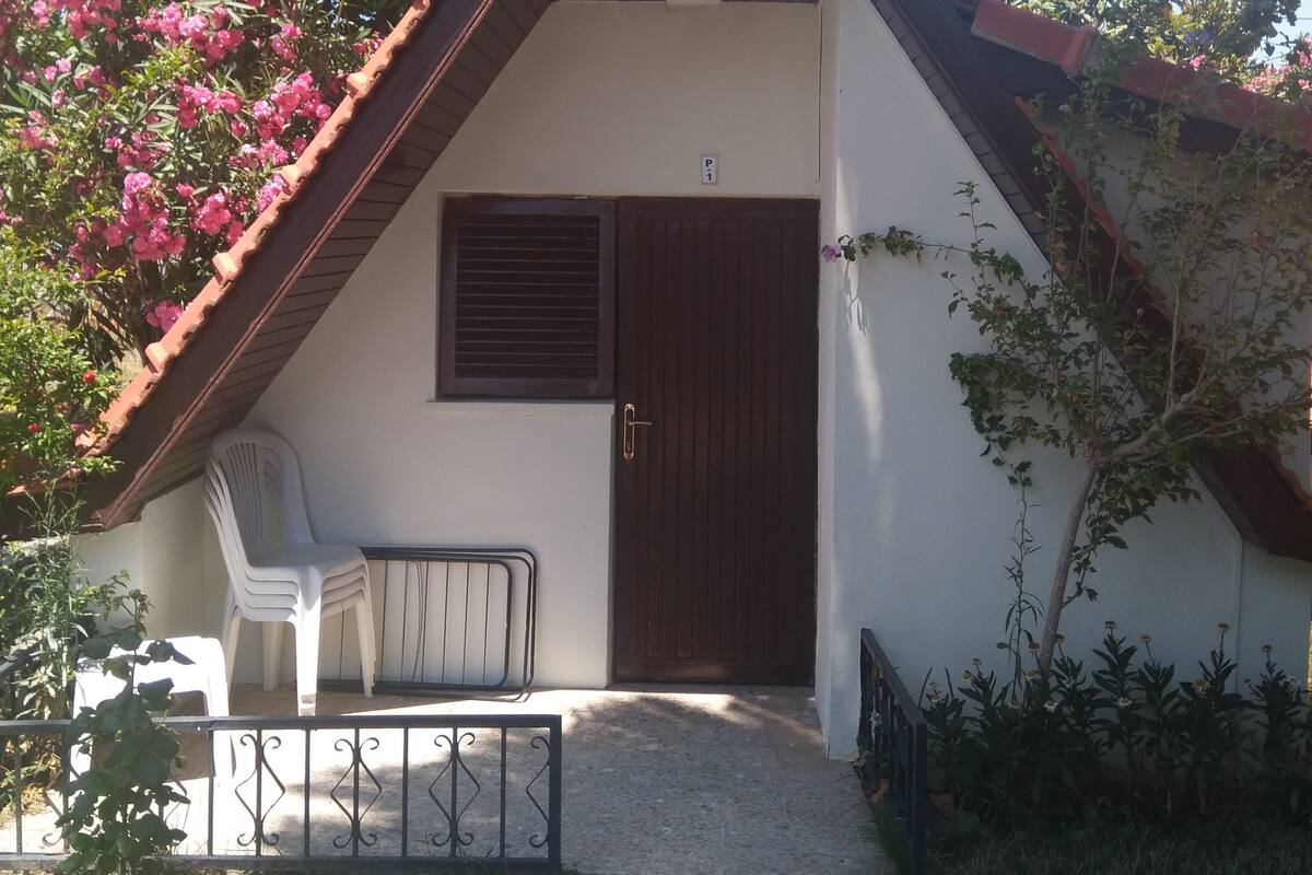 A small, single-story house with a sloped roof, a front door, and a small porch area with a chair.