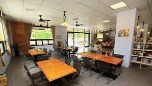 A spacious dining area with multiple tables and chairs, large windows with a view of greenery, ceiling fans, and a bookshelf with various items. - Fu Chuan Garden Homestay (Nanzhuang)