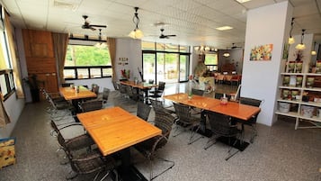 A spacious dining area with multiple tables and chairs, large windows with a view of greenery, ceiling fans, and a bookshelf with various items.