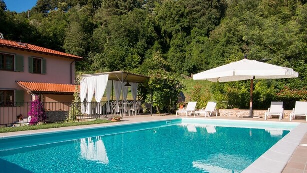 A swimming pool with clear blue water, surrounded by lounge chairs and umbrellas, and a house with a red-tiled roof in the background. - Il Poggiolo (Camaiore)