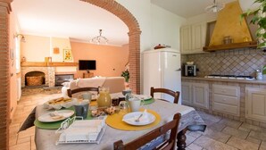 A dining area with a brick archway, a fireplace, and a kitchen with white cabinets and a yellow range hood. - Il Poggiolo (Camaiore)