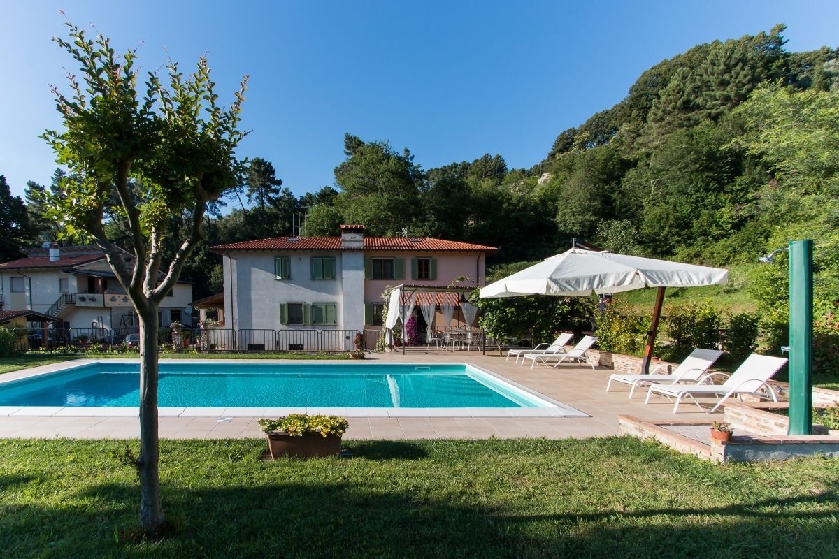 A swimming pool with lounge chairs and umbrellas, surrounded by a grassy area and a building with a red roof.