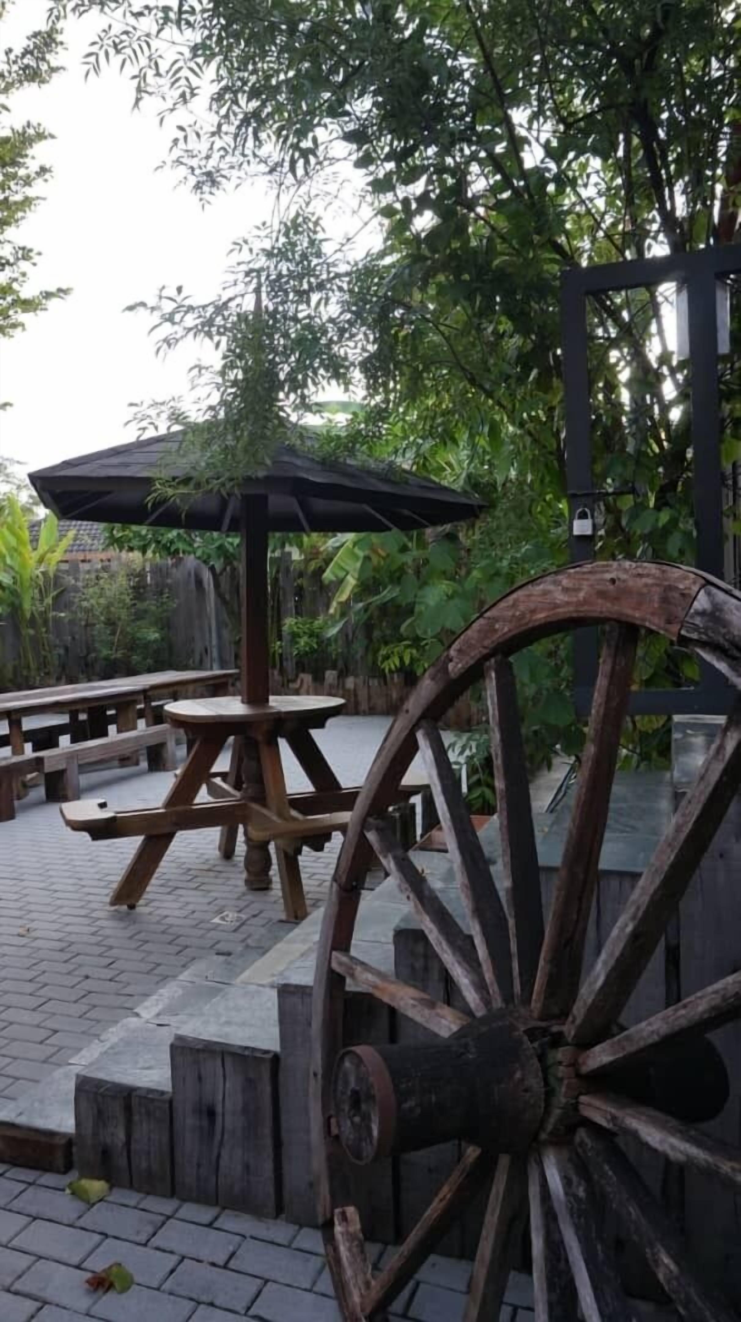 A rustic outdoor seating area with wooden tables and benches, surrounded by lush greenery and a large wooden wheel structure.