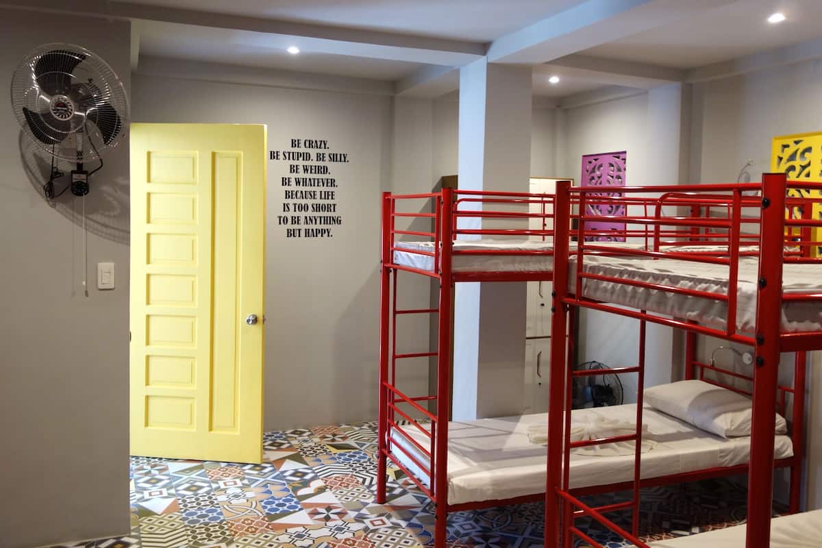 Una habitación con literas, un ventilador, una frase motivacional en la pared y un suelo estampado.
