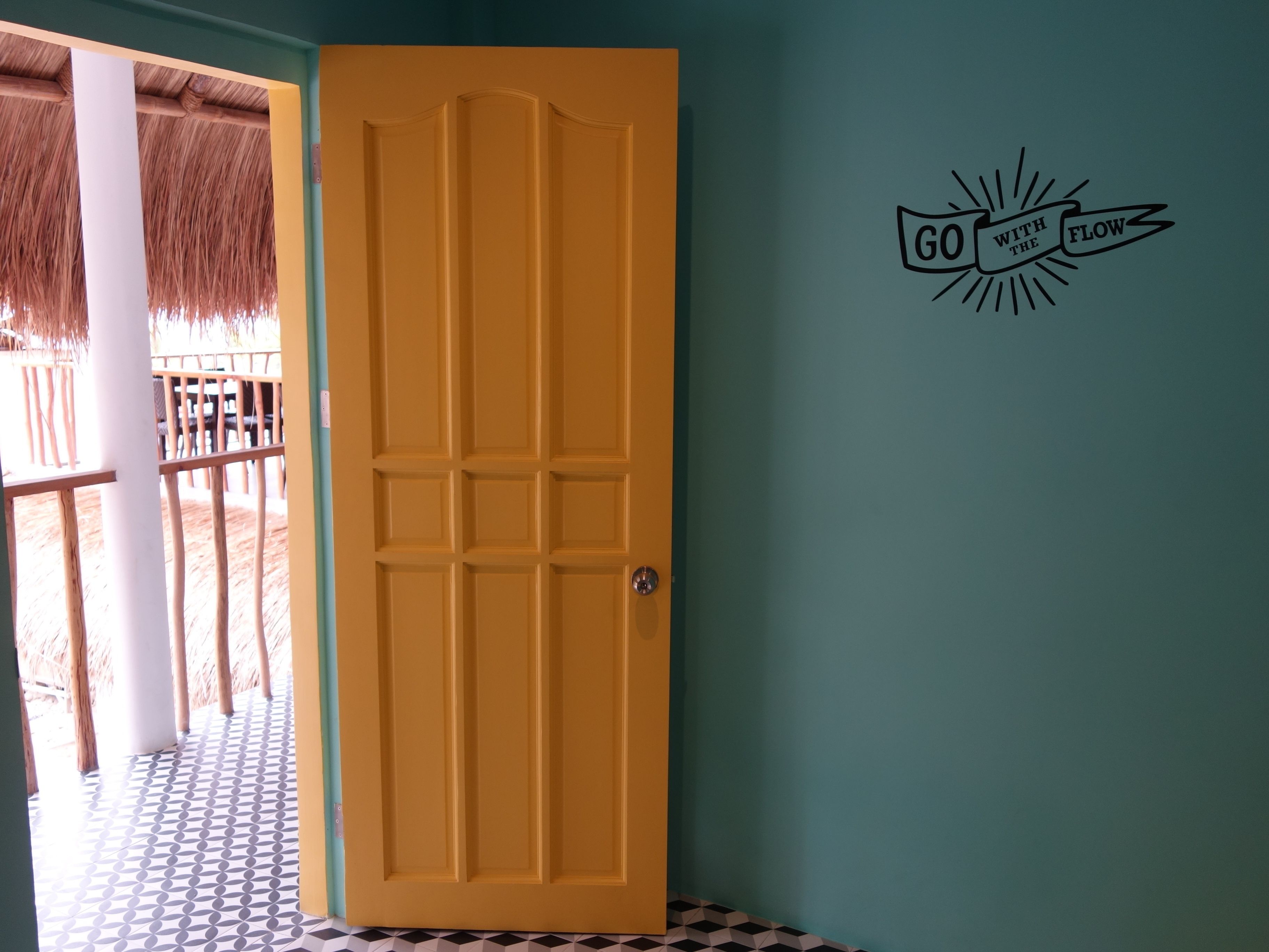 Une porte jaune ornée d’un panneau décoratif, une structure à toit de chaume et un mur sur lequel est inscrit le message « Laissez-vous porter par le courant ».