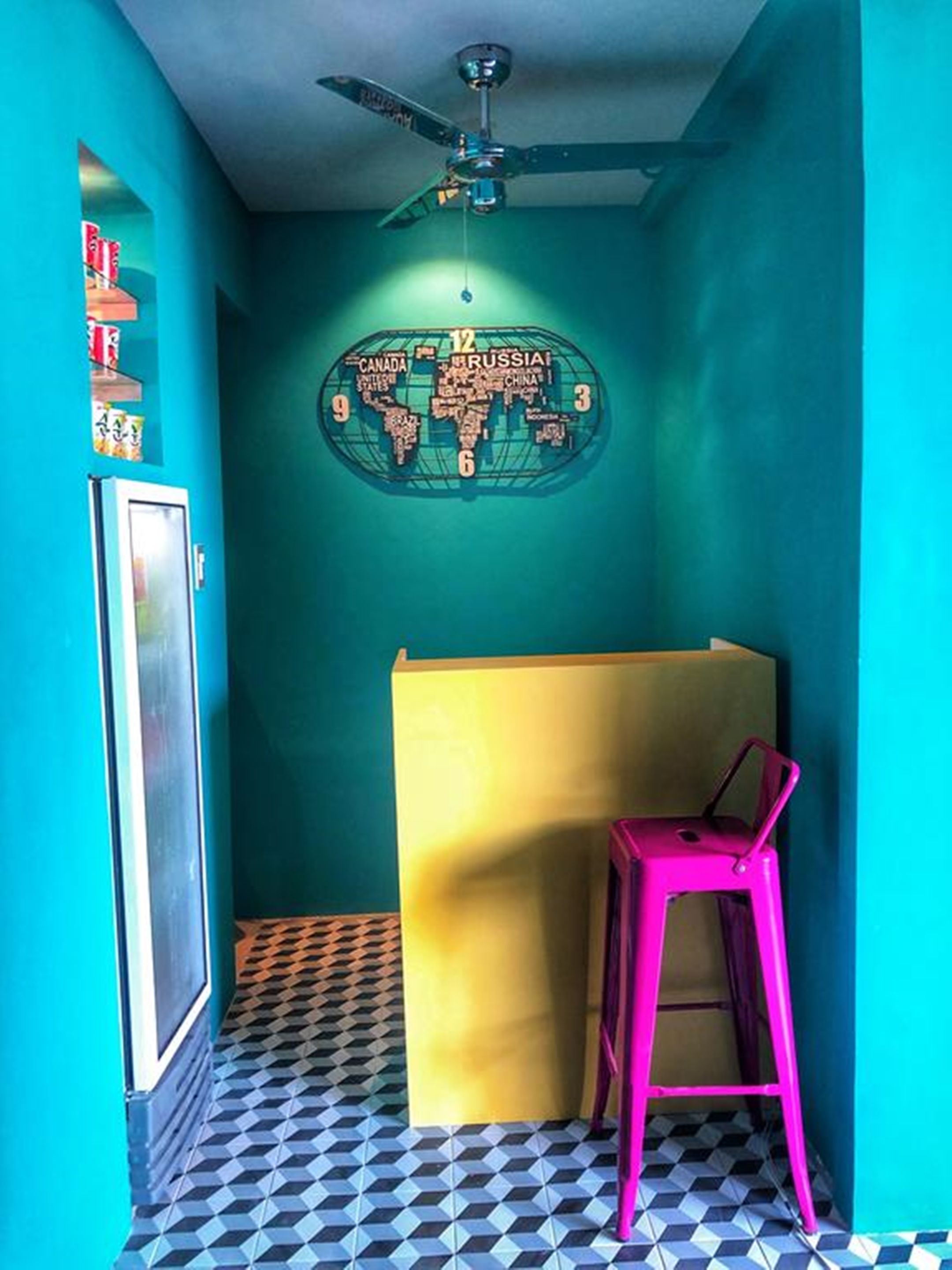 A room with teal walls, a black and white checkered floor, a ceiling fan, a map on the wall, a yellow partition, and a pink chair.