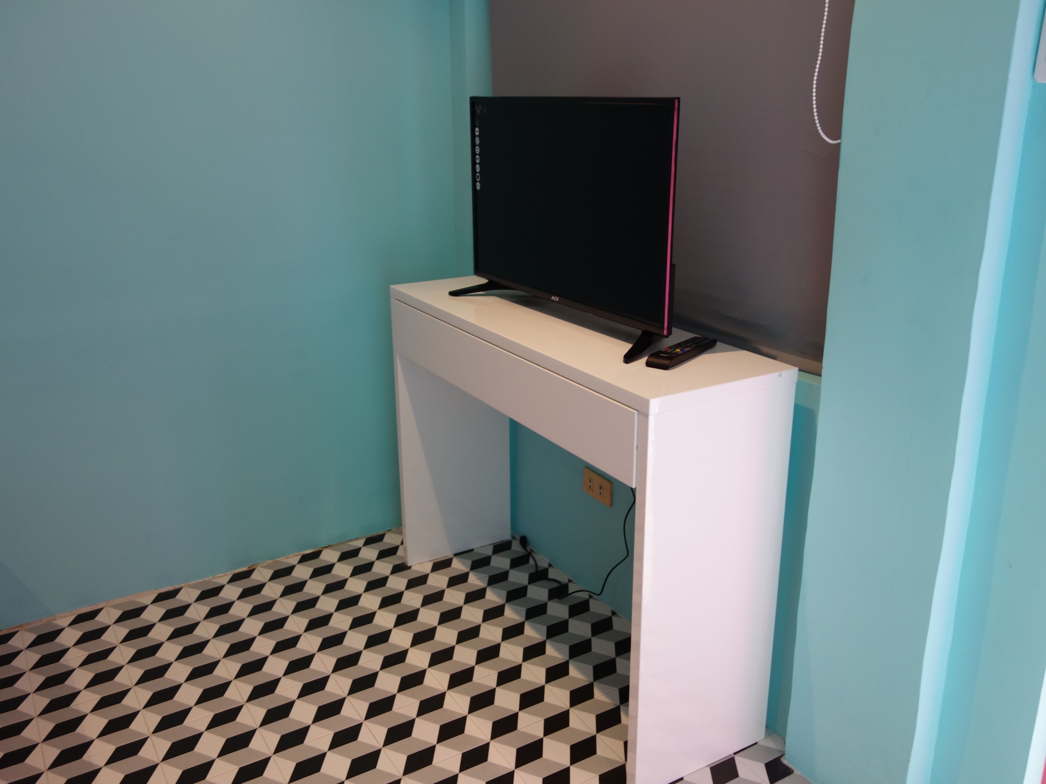A small room with a white desk, a black monitor, and a remote control on it. The floor has a black and white checkered pattern.