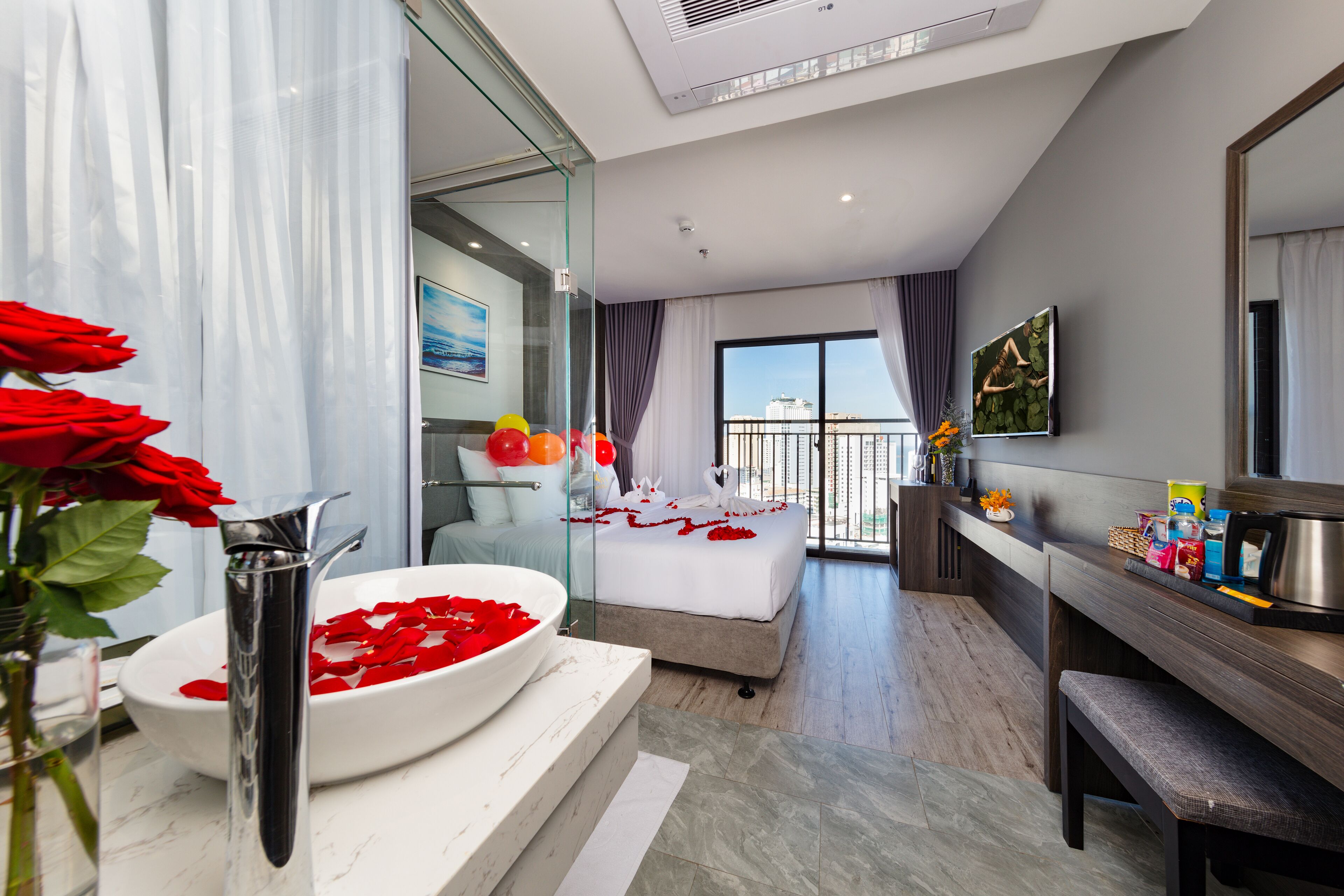 A modern hotel room with a large bed, a bathroom with a white sink and rose petals, a balcony with a view, and a wooden vanity.