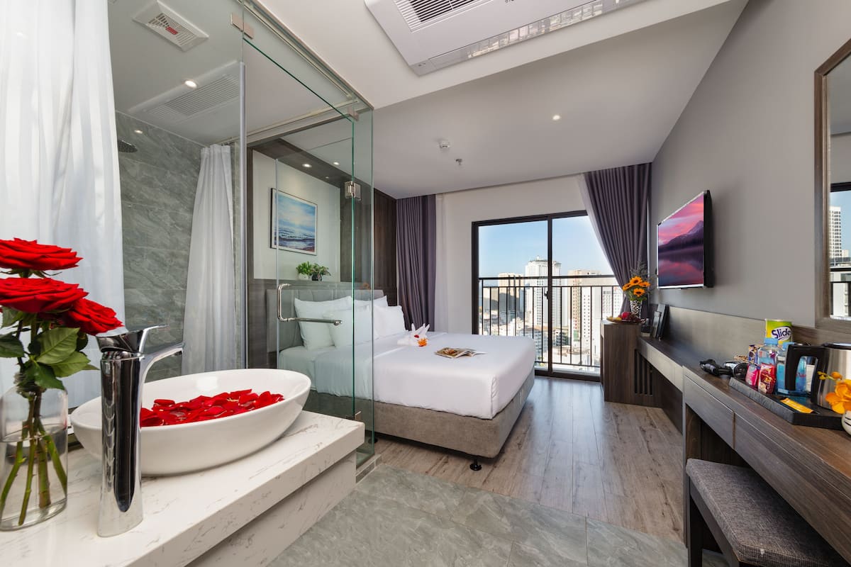 Une chambre d’hôtel moderne dotée d’un grand lit, d’une douche à parois de verre, d’un lavabo blanc et d’un balcon offrant une vue sur la ville.