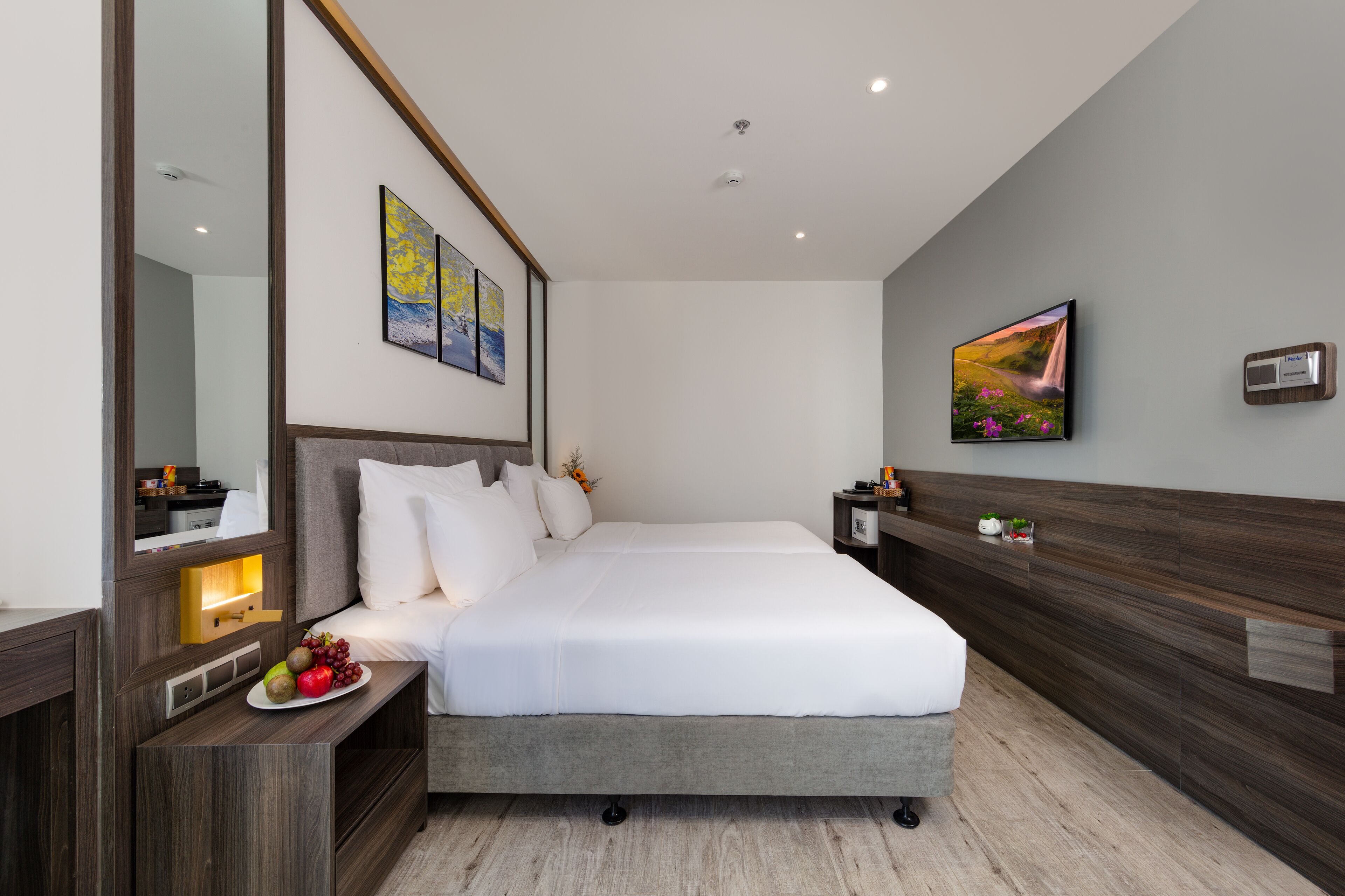 Une chambre d’hôtel moderne avec un grand lit, une tête de lit en bois, des tables de chevet et des œuvres d’art accrochées aux murs.