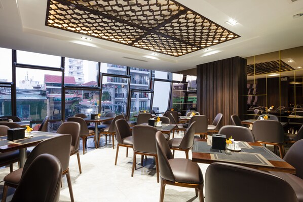 A modern hotel dining area with multiple tables and chairs, large windows offering city views, and a decorative ceiling.