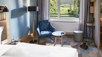 Une chambre d’hôtel avec un lit, un fauteuil bleu, une petite table ronde, une lampe sur pied, une fenêtre donnant sur un paysage verdoyant et des montagnes, et un porte-chaussures.