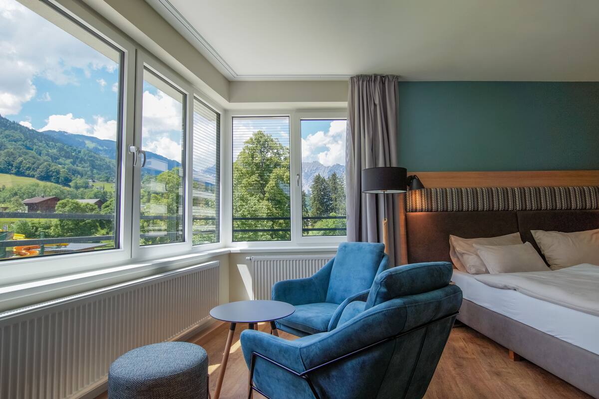 A hotel room with a bed, a blue armchair, a small round table, and large windows offering a mountain view.