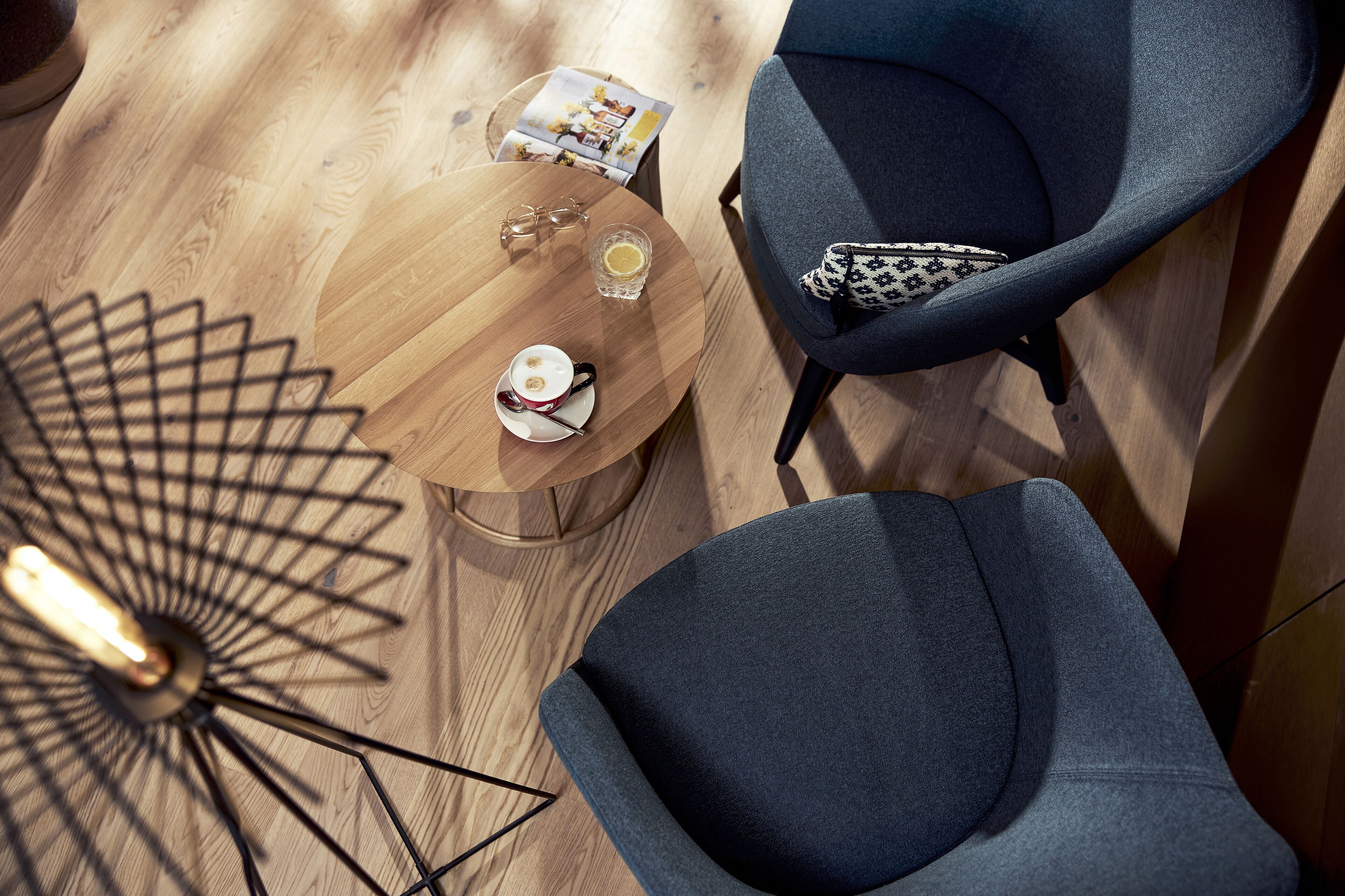 A modern room with a wooden floor, a round wooden table with a glass, a cup, and a magazine, and two grey armchairs with patterned cushions.