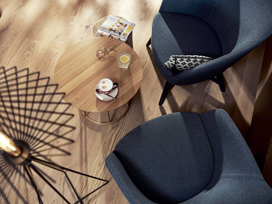 A modern room with a wooden floor, a round wooden table with a glass, a cup, and a magazine, and two grey armchairs with patterned cushions.