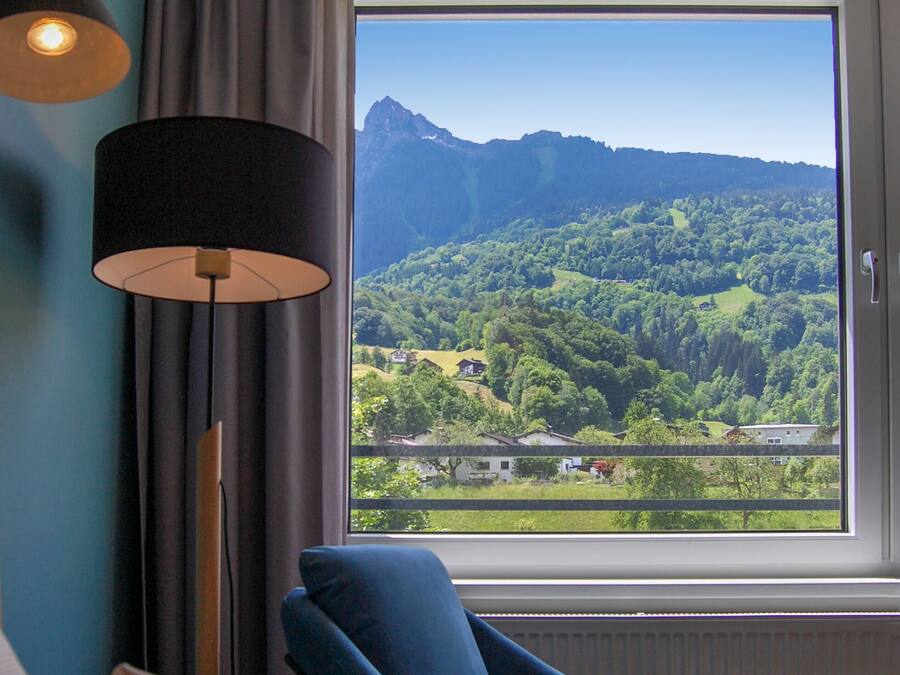 A room with a large window offering a view of a mountainous landscape. There is a blue armchair and a floor lamp.