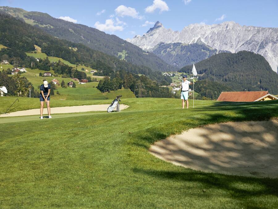 Un campo de golf con montañas al fondo, un búnker de arena y un mástil con bandera.