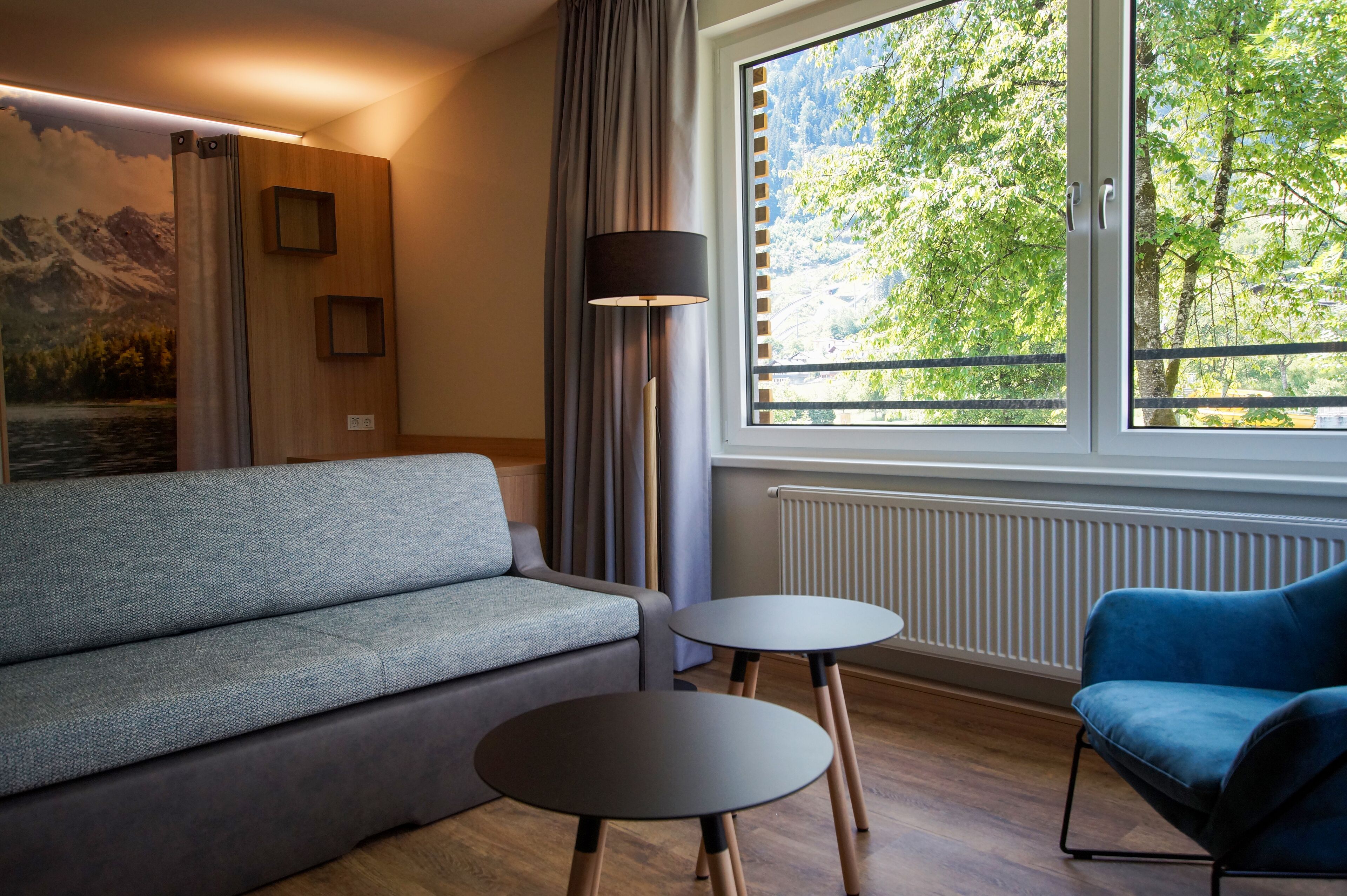 A modern living room with a grey sofa, two round tables, a floor lamp, and a window with a view of trees.