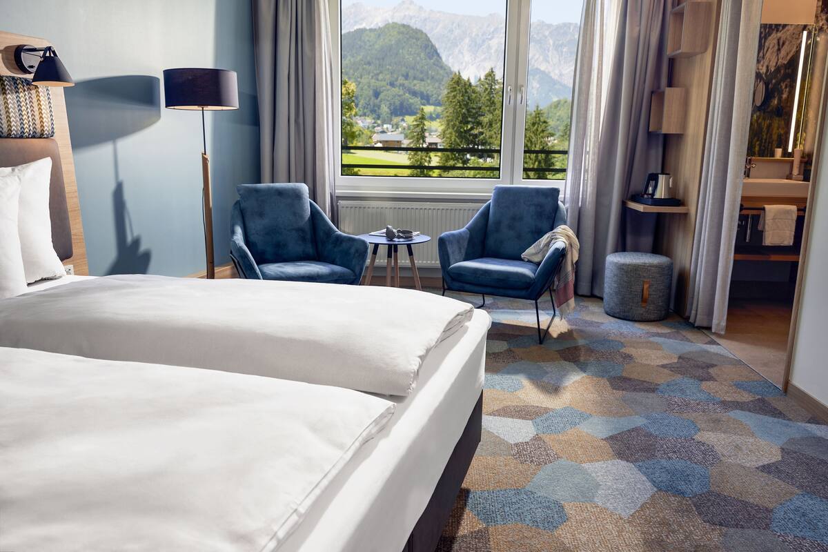 A hotel room with a bed, two armchairs, a small table, a view of mountains, and a bathroom with a mirror and sink.