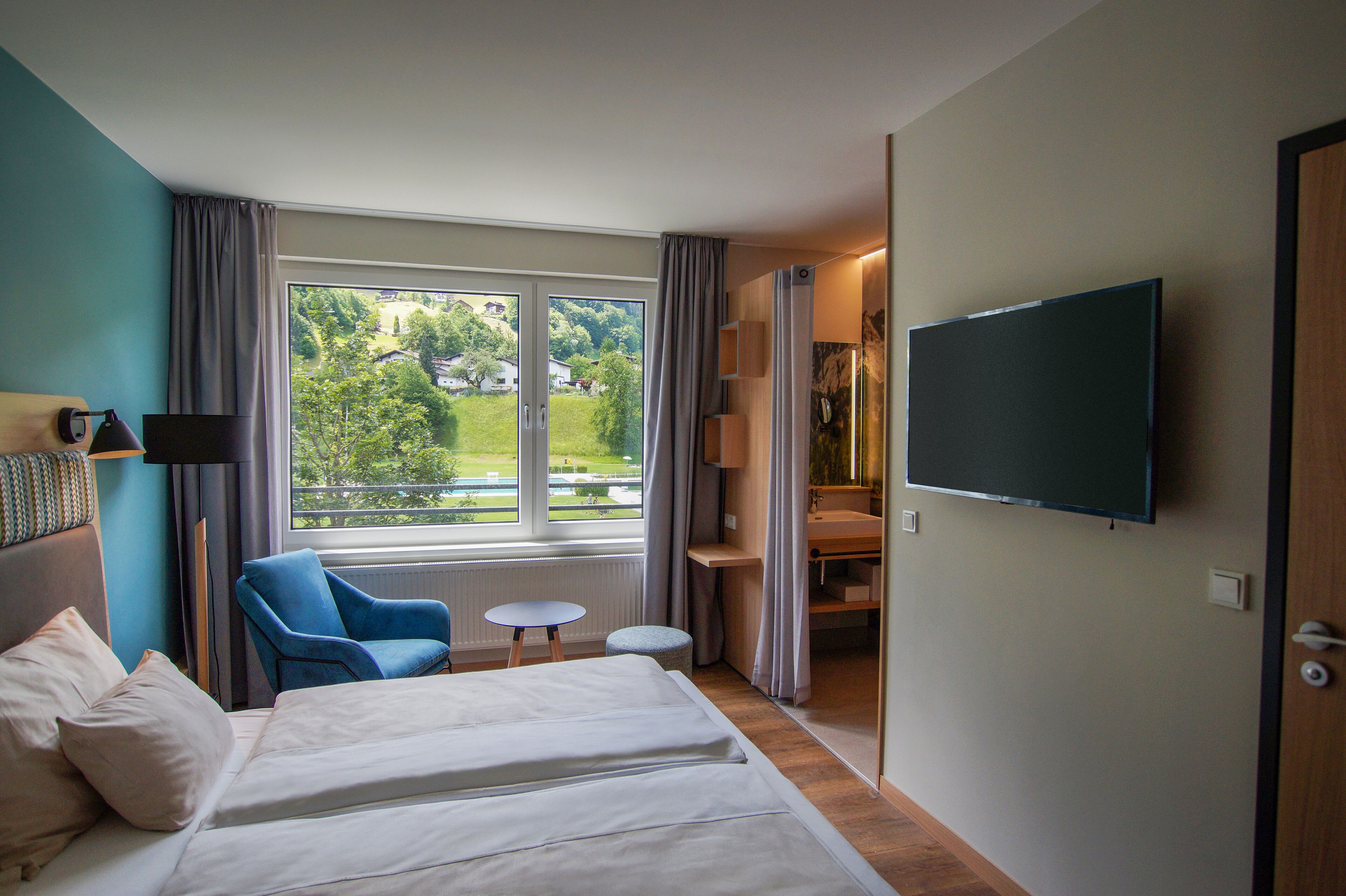 A hotel room with a bed, a blue armchair, a small table, a TV, and a view of greenery outside.