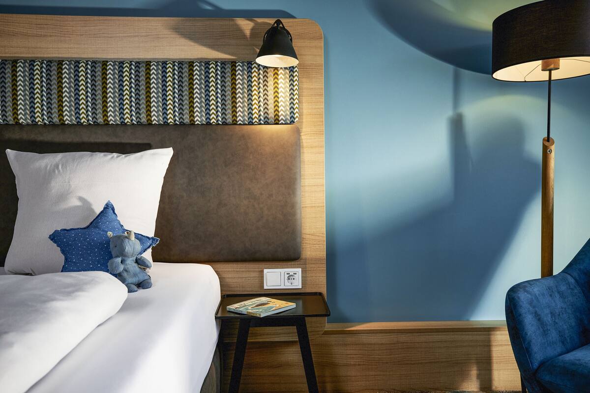 A neatly made bed with a pillow, a bedside lamp, and a small table with a magazine.