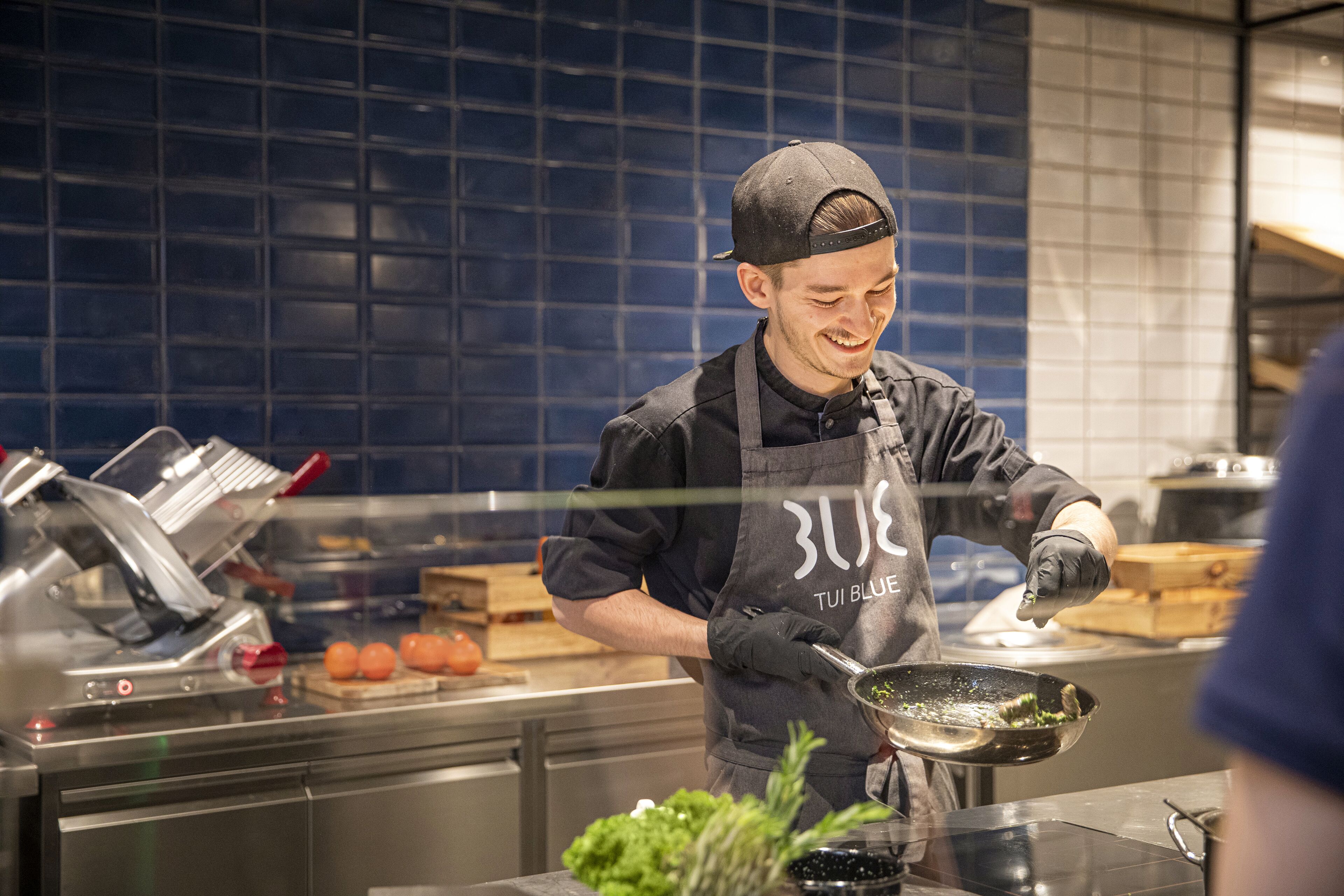 A chef in a kitchen with a blue tiled backsplash, preparing food on a stove.