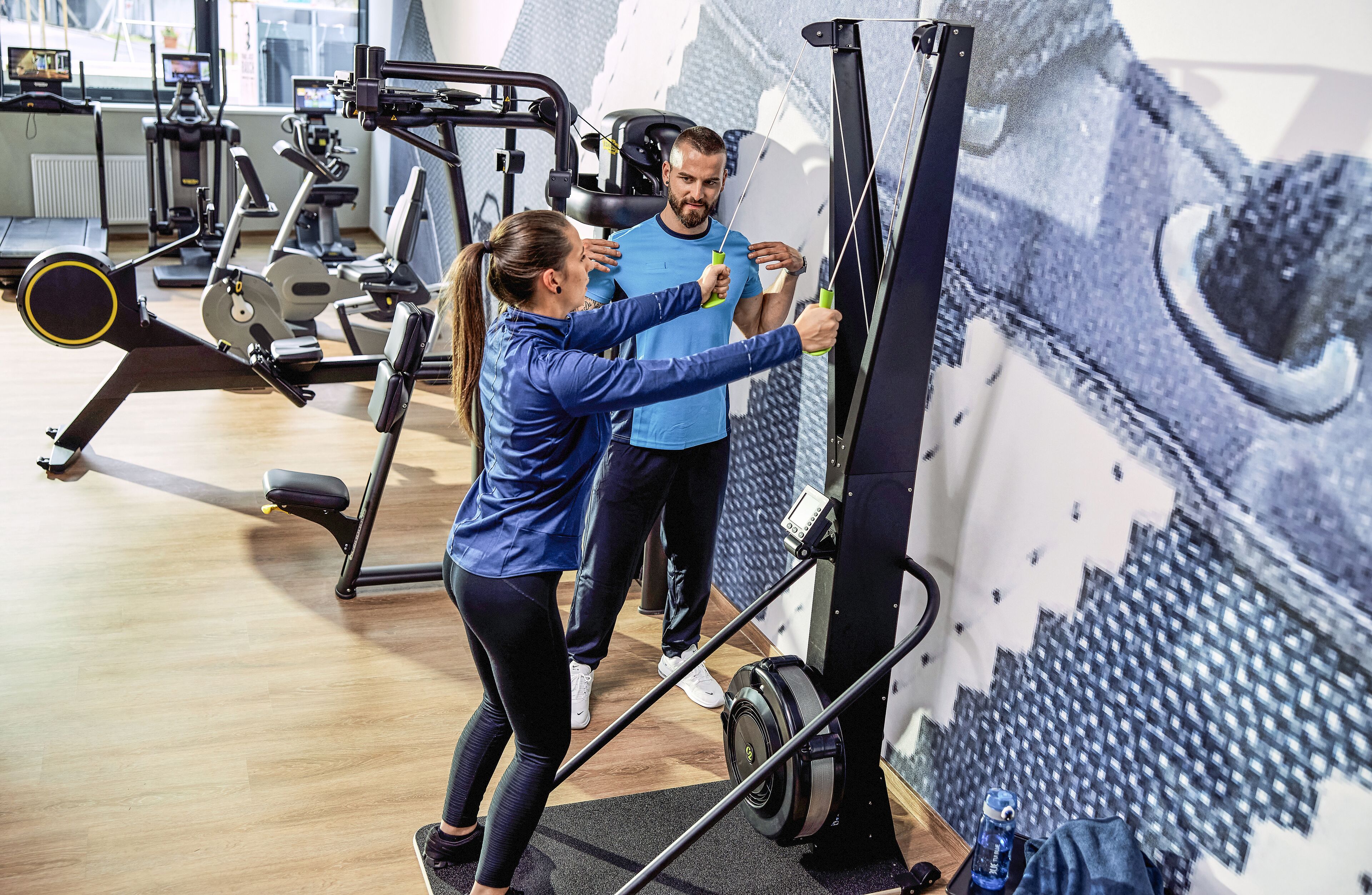Un gimnasio con equipo de ejercicio, una pared con un mural grande y una persona usando una máquina de remo.