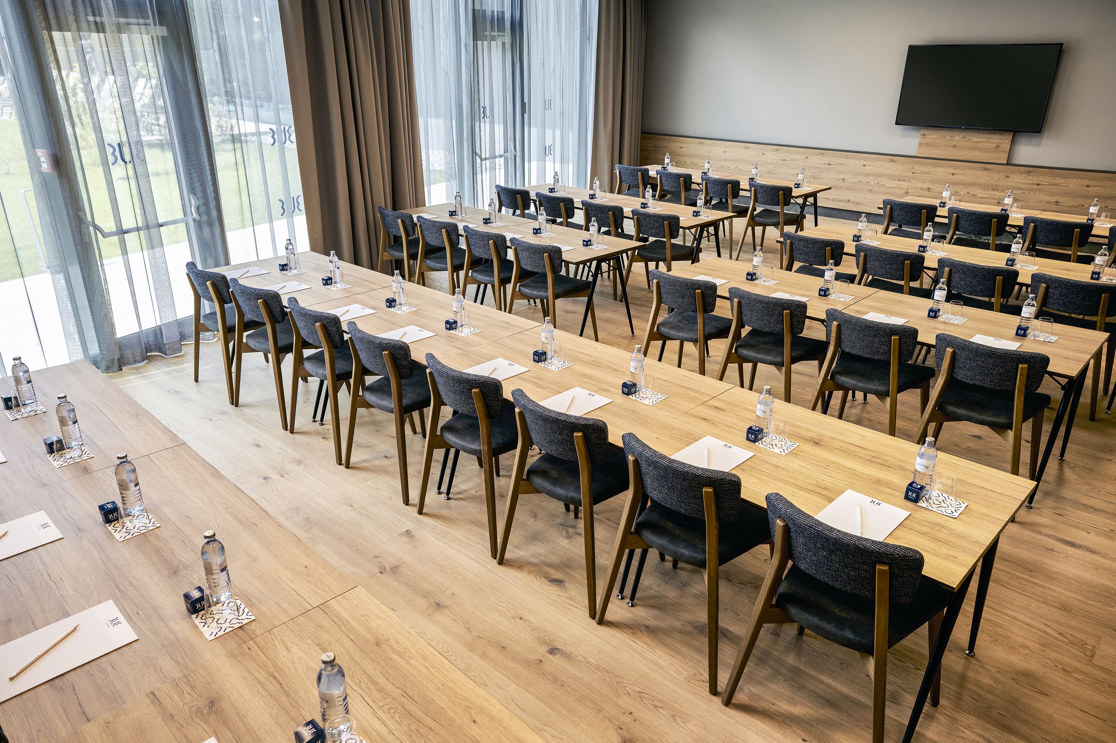 Une salle de conférence dotée de plusieurs tables et chaises, de grandes fenêtres avec des rideaux et d’un téléviseur à écran plat fixé au mur.