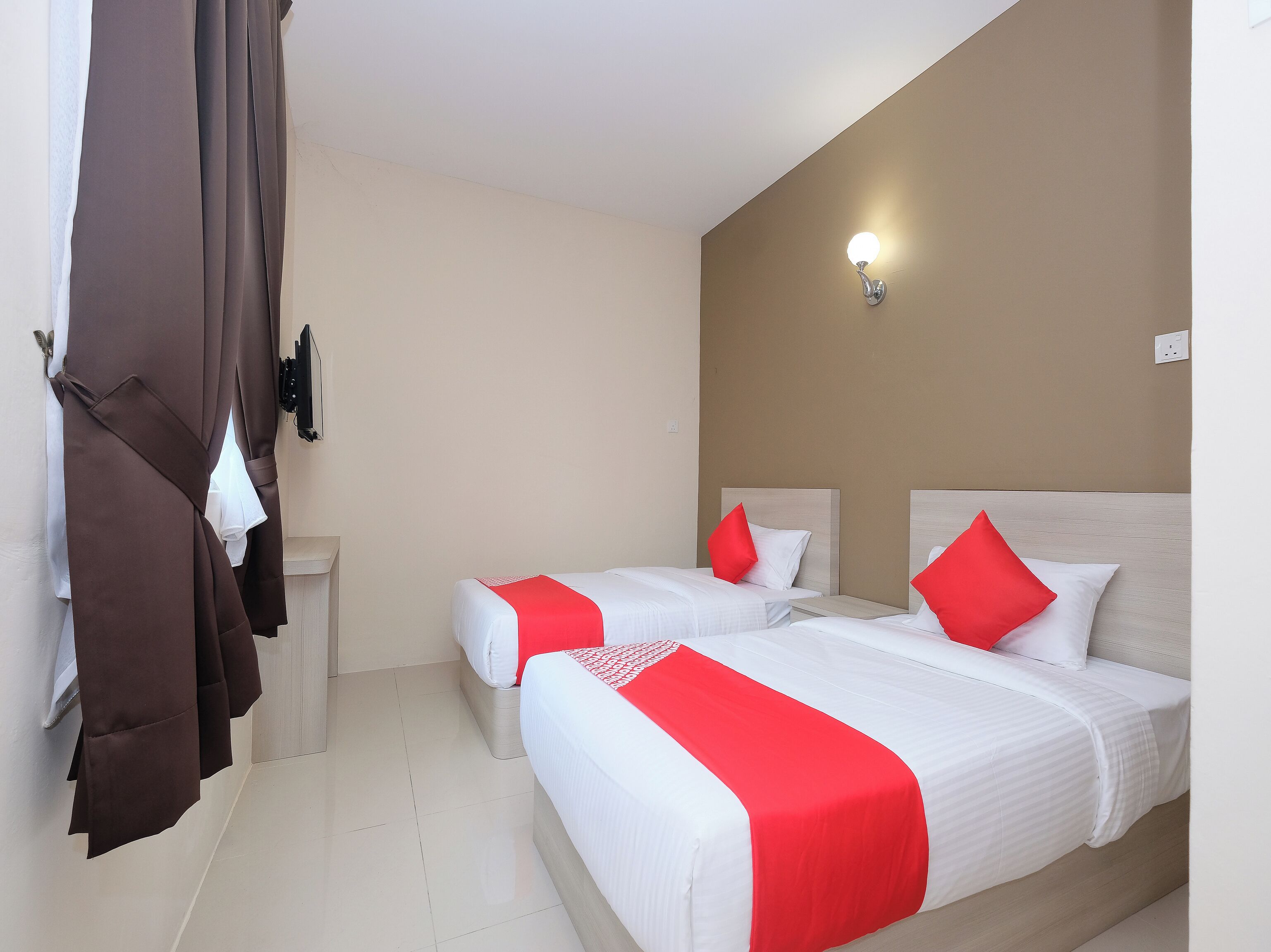 A hotel room with two beds, each with white bedding and red accents, a light-colored wall, and a ceiling light fixture.