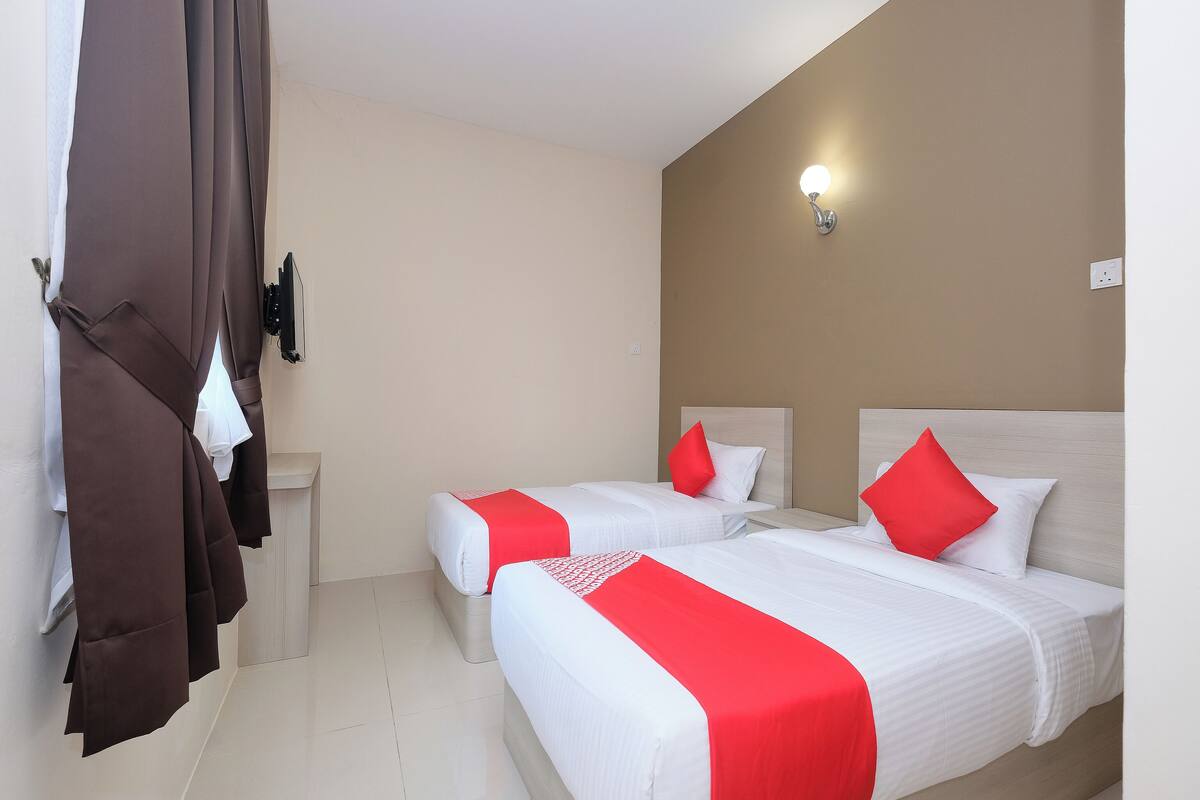 A hotel room with two beds, each with white bedding and red accents, a light-colored wall, and a ceiling light fixture.