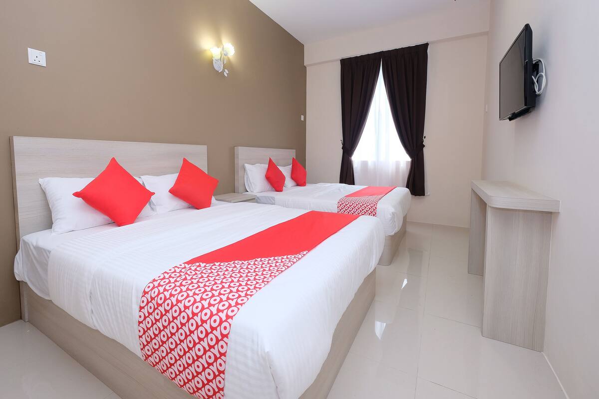 A hotel room with two beds, red pillows, a red and white patterned bed runner, a flat-screen TV mounted on the wall, and a window with dark curtains.