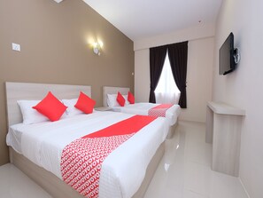 A hotel room with two beds, red pillows, a red and white patterned bed runner, a flat-screen TV mounted on the wall, and a window with dark curtains.