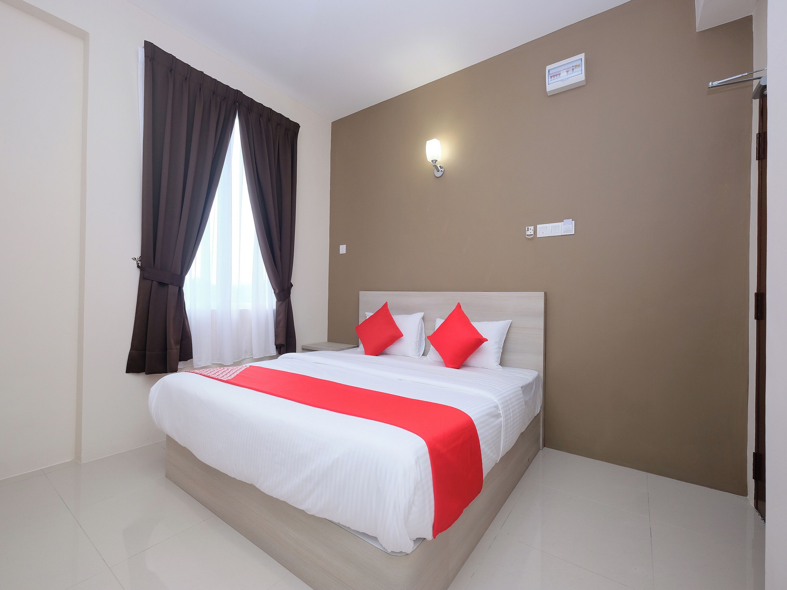 A hotel room with a bed, curtains, a wall-mounted light, and a door.