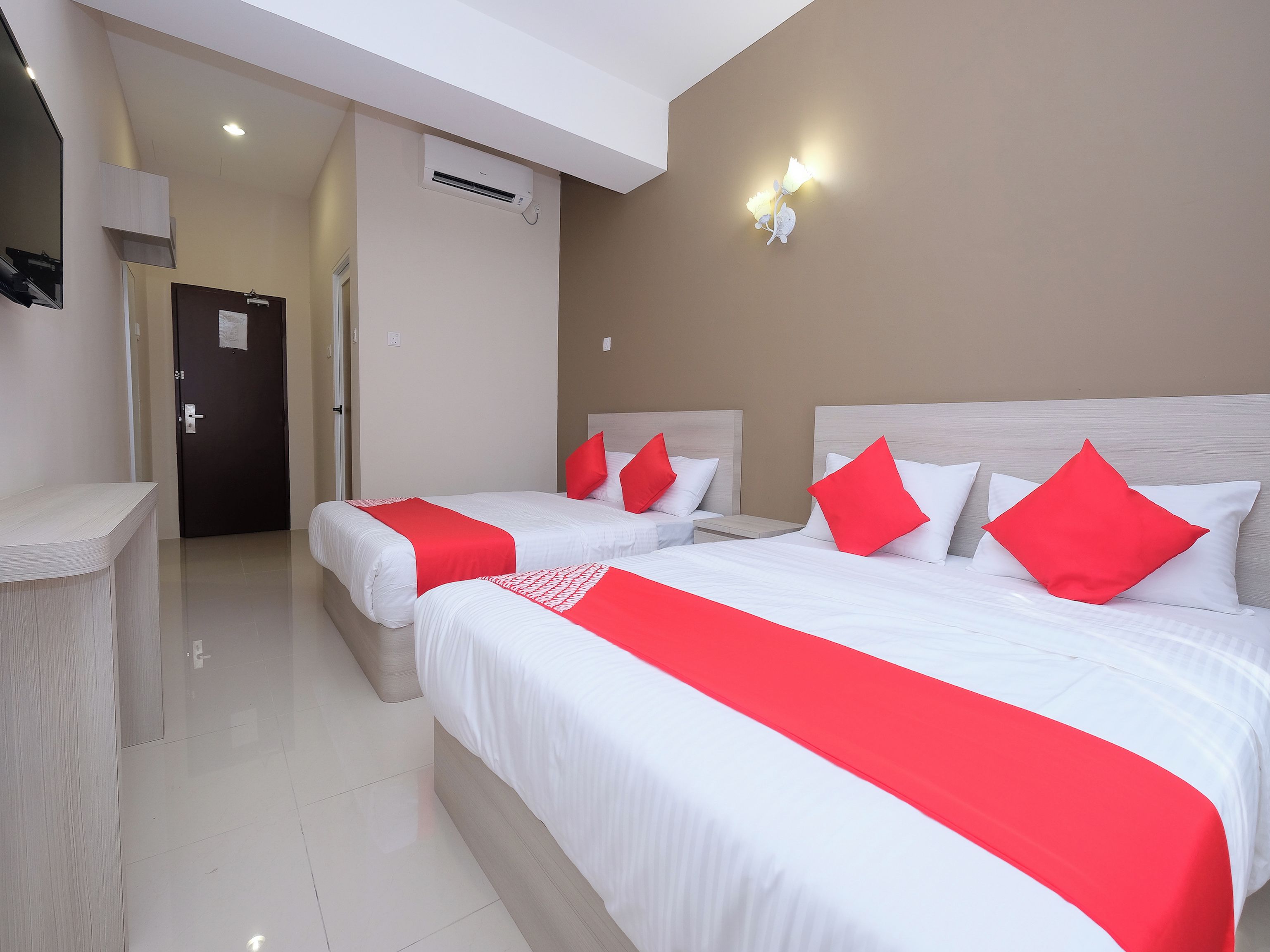 A hotel room with two beds, each with white bedding and red accents, a desk, a door, and an air conditioning unit.
