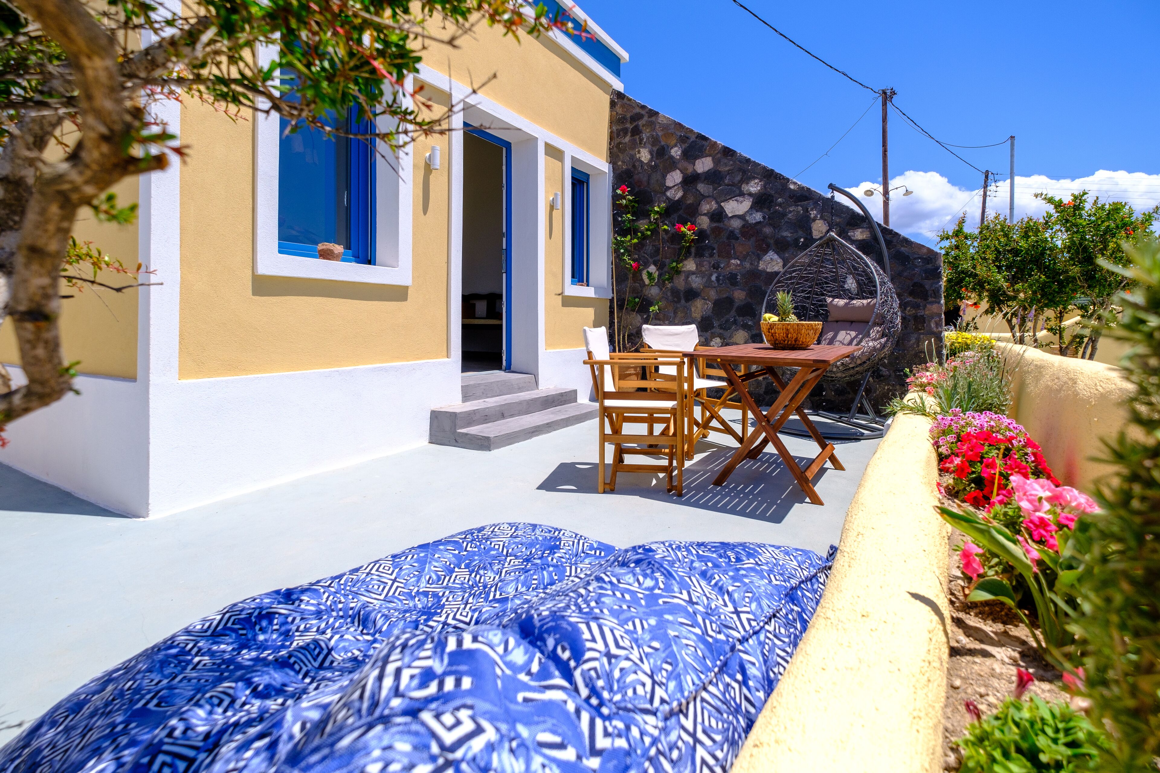 Un espacio al aire libre acogedor con una mesa, sillas y una mecedora de mimbre, además de un cojín azul estampado.
