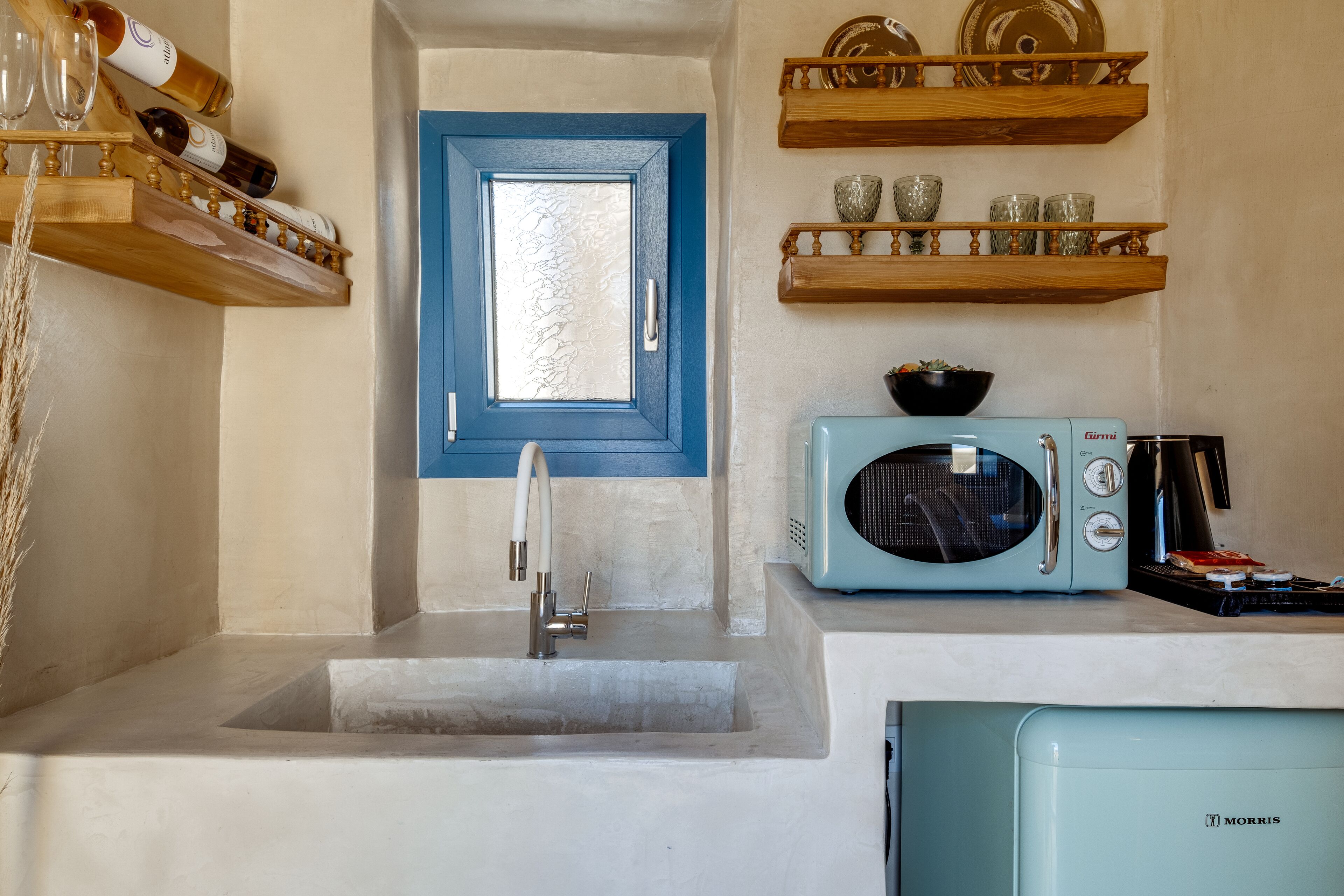 A compact kitchen with a blue door, a white sink, a vintage Morris microwave, and wooden shelves above holding glassware and dishes.