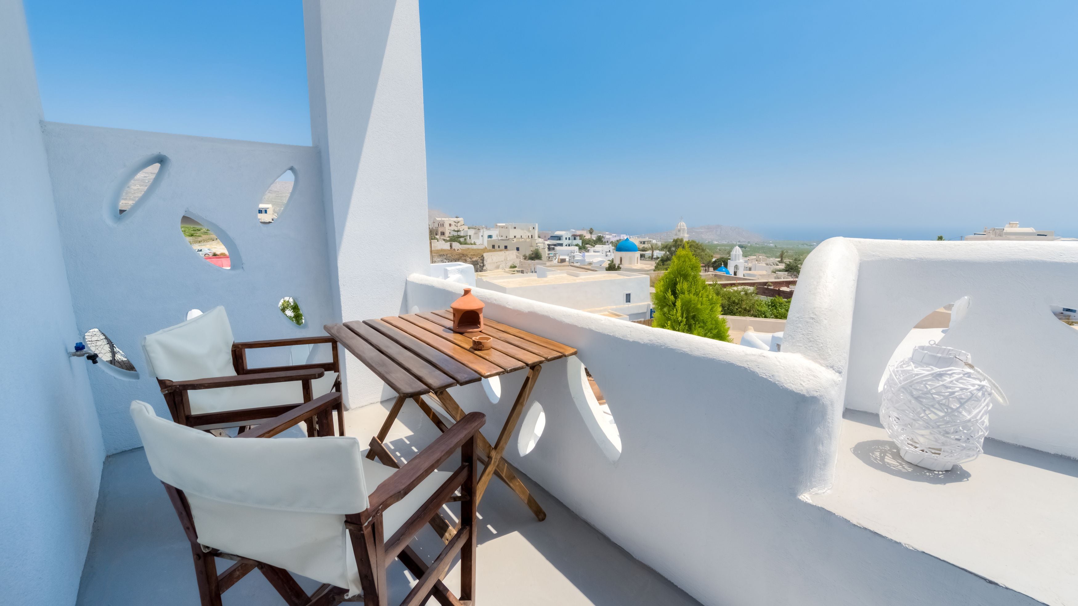 A balcony with a wooden table and two white chairs, overlooking a coastal town with white buildings and a clear blue sky.