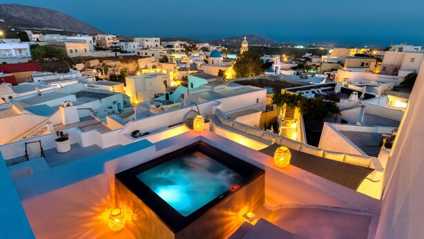 A rooftop with a hot tub and ambient lighting, overlooking a coastal town with white buildings and a mountain in the background. - Pasithea Luxury Suites (Santorini)