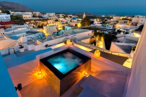 Una azotea con jacuzzi e iluminación ambiental, con vistas a un pueblo costero de edificios blancos y una montaña al fondo.