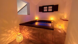 A spa room with a wooden bathtub, a small window, and a wall-mounted shelf with candles. - Pasithea Luxury Suites (Santorini)