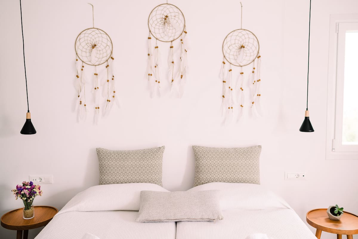 Quarto bem arrumado com cama, almofadas estampadas, mesa de cabeceira com um vaso de flores e três sonhadores pendurados.