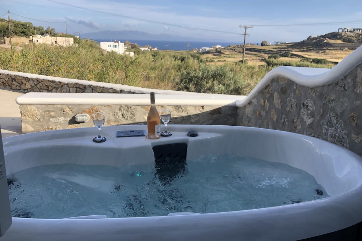 Uma banheira de hidromassagem com uma garrafa de vinho e duas taças, com vista para uma paisagem pitoresca com edifícios e colinas.