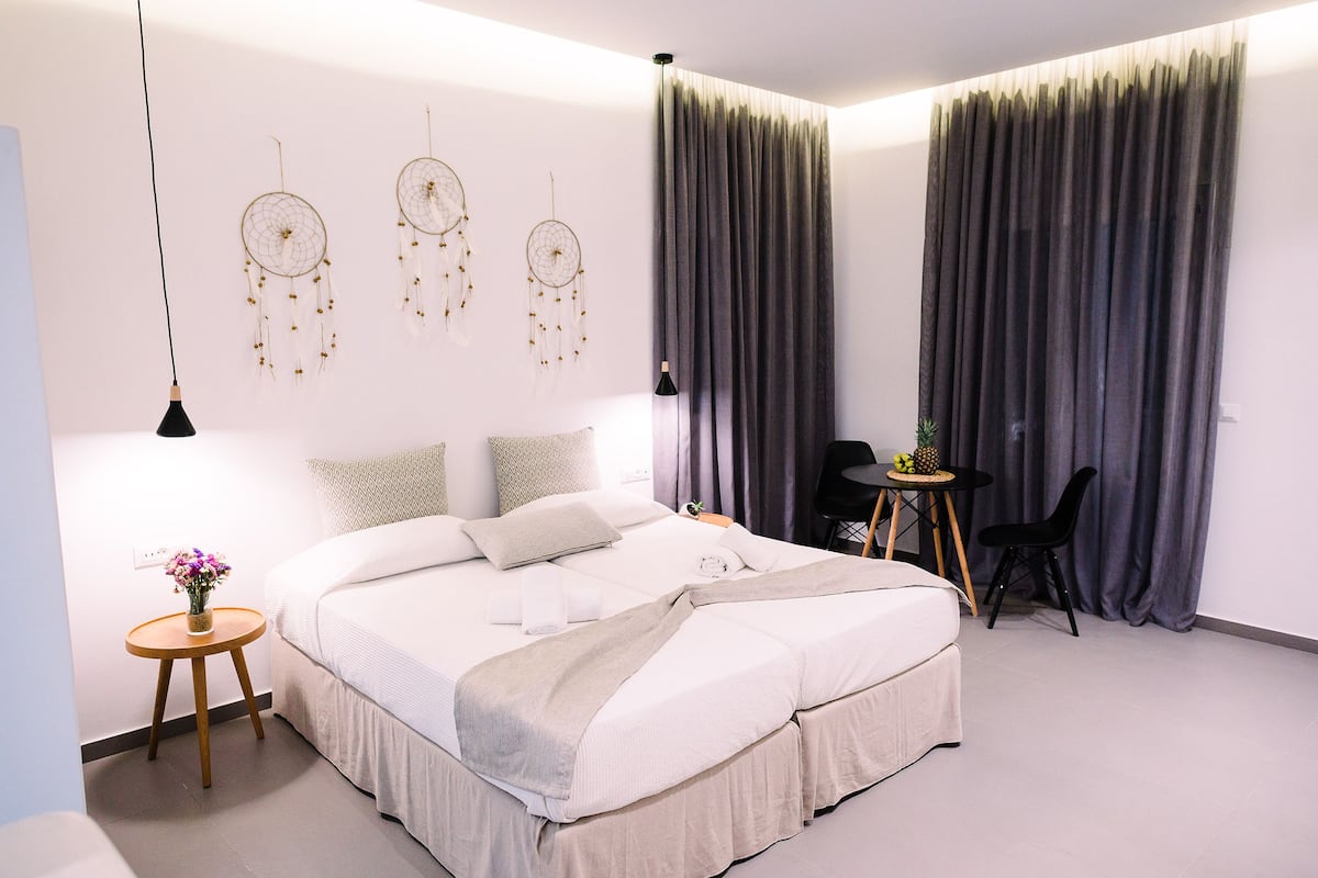 Um quarto moderno com uma cama grande, uma mesa de cabeceira com abajur, uma pequena mesa redonda com uma cadeira e um sonho-pescador fixado na parede.