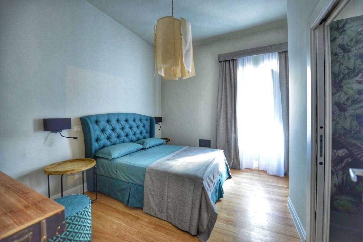 Une chambre avec un grand lit, une table de chevet, un luminaire suspendu et une vue sur l’extérieur par une fenêtre ornée de rideaux.