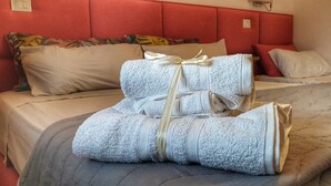Una cama bien tendida con colcha gris y toalla blanca con listón amarillo, en una habitación con cabecera roja y varios almohadones.