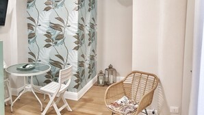 Una habitación con una mesa redonda de vidrio, dos sillas blancas, una silla de mimbre, un panel decorativo con motivos de hojas y una lámpara colgante.