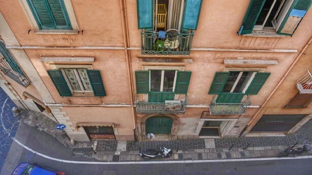 Un edificio de varios pisos con contraventanas verdes, un balcón donde se ve una bicicleta y una entrada a nivel de calle.