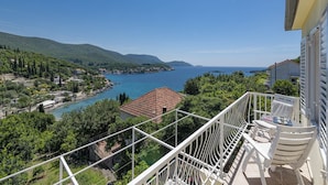 Ein Balkon mit weißen Geländern, der auf eine Küstenstadt, das klare blaue Meer und hügelige grüne Landschaften blickt.