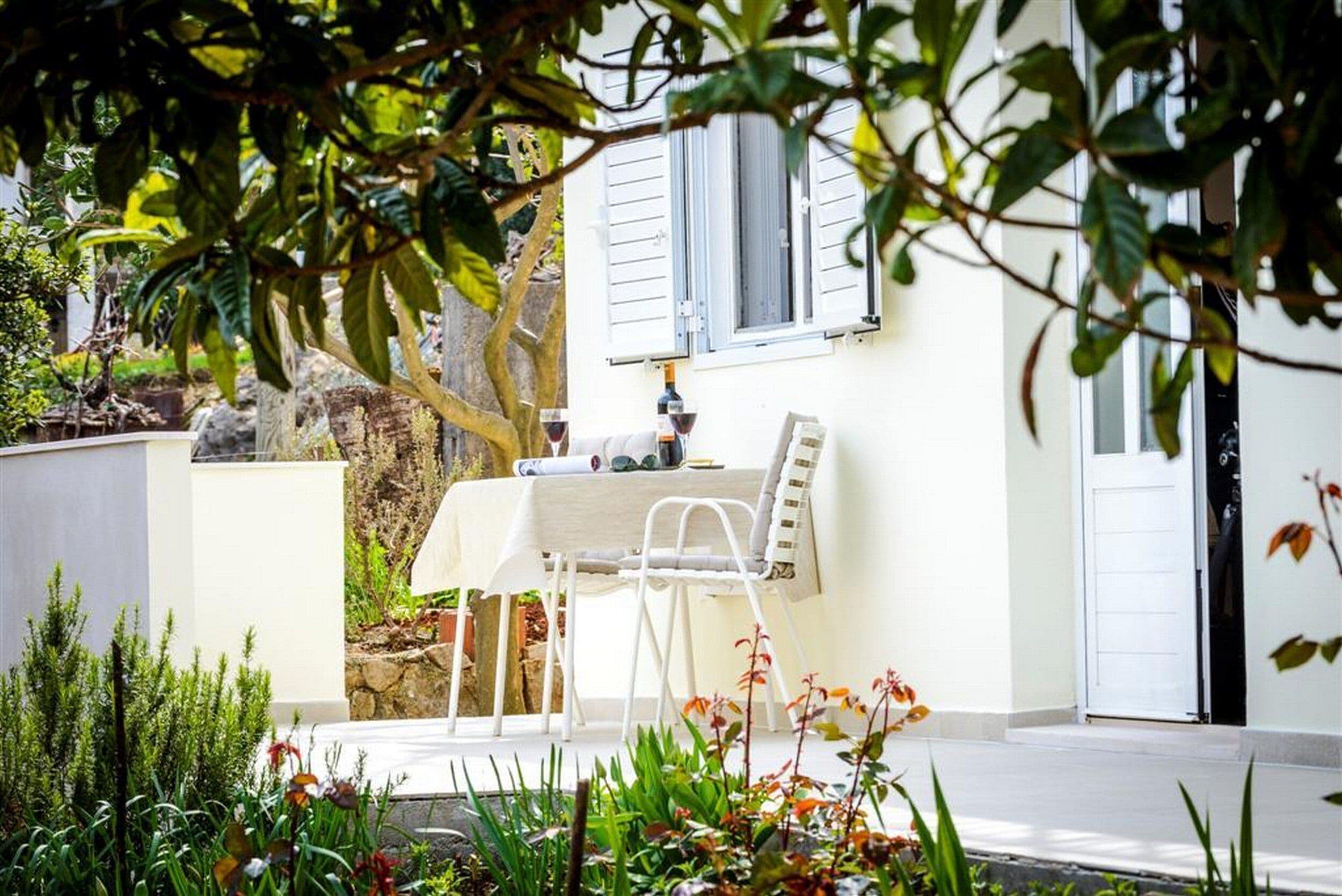 A white table with a chair outside a house, a bottle of wine, and a window with shutters.